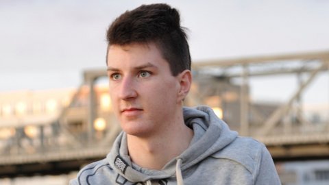 Junger Mann mit dunklen Haaren trägt eine graue Kapuzenjacke, im Hintergrund eine Stahlbrücke und Gebäude bei Tageslicht.
