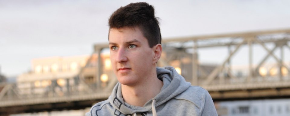Junger Mann mit dunklen Haaren trägt eine graue Kapuzenjacke, im Hintergrund eine Stahlbrücke und Gebäude bei Tageslicht.