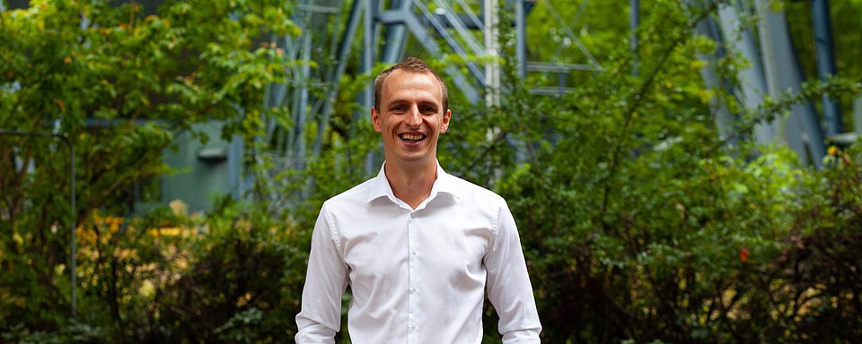 Mann in weißem Hemd steht vor grünen Büschen und einer grauen Stahlstruktur im Hintergrund