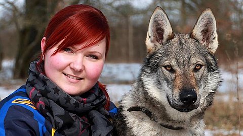 Großer grauer Hund mit aufrechten Ohren sitzt neben einer rothaarigen Frau im blauen Jacke und schwarzem Schal, Hintergrund mit Bäumen und Schnee