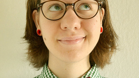 Frau mit schulterlangen braunen Haaren, trägt eine dunkle Brille, grün-weiß kariertes Hemd und rote Ohrringe, vor heller Wand