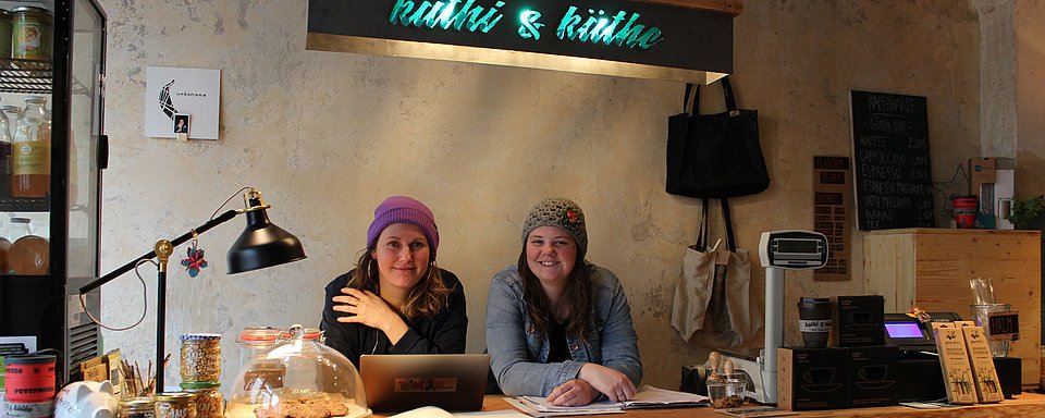 Zwei Frauen sitzen an einem Holztisch mit Laptop, Papieren und einer Lampe in einem gemütlichen Café mit rustikaler Einrichtung.