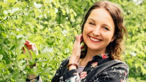 Erlebnisse in der Natur von intogreen.events Frau mit lockigem Haar trägt schwarzen Pullover mit Blumenmuster, steht im Grünen und berührt Blätter eines Baums