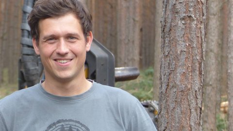 Junger Mann mit kurzem braunem Haar, grauem T-Shirt mit rundem schwarzen Muster, steht vor einem Wald mit vielen hohen Bäumen.