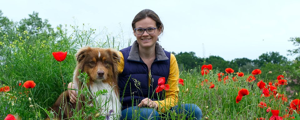 Frau mit dunkelblauer Weste und gelbem Langarmshirt sitzt in einem Mohnblumenfeld und hält eine braune Papiertüte hoch.